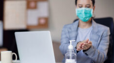 woman with mask on in front of laptop, sanitising her hands