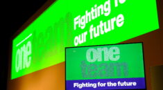 Health conference stage background: a green themed graphic which says One Team Fighting for out future, a TV in front of the background shows a similar graphic with the same words.