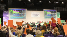 Delegates to UNISON's women's conference raising organ voting cards, photographed from the back of the hall so the conference set is visible in the background