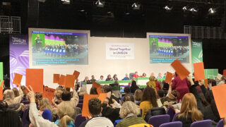 Delegates to UNISON's women's conference raising organ voting cards, photographed from the back of the hall so the conference set is visible in the background