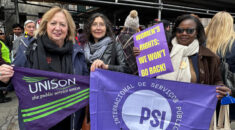 Christina McAnea standing at a rally with two women, holding trade union flags, including a UNISON one, with other women in the background
