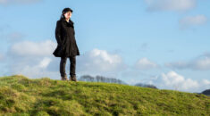 Image of a young woman standing on the top of a green hill, with blue sky behind her