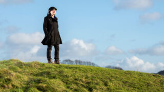 Image of a young woman standing on the top of a green hill, with blue sky behind her