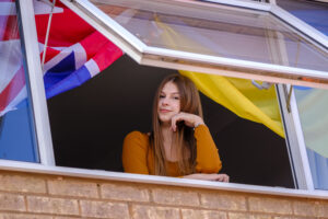 Vlada looking out of her bedroom with a Ukrainian flag and a union flag (union jack) hanging next to her