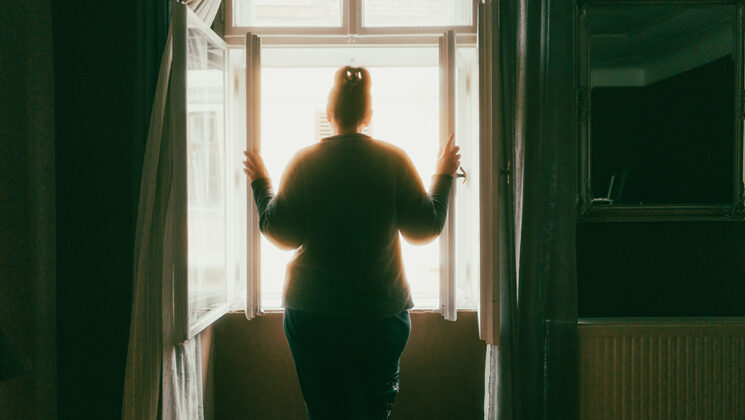 Color image depicting the rear view of a lonely woman looking at the street from her bedroom window. She is rendered an almost total silhouette due to the light flooding into the room from the window.