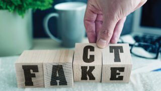 Four wooden blocks with a hand turning the third and fourth so that the letters spell out 'fact' instead of 'fake'.