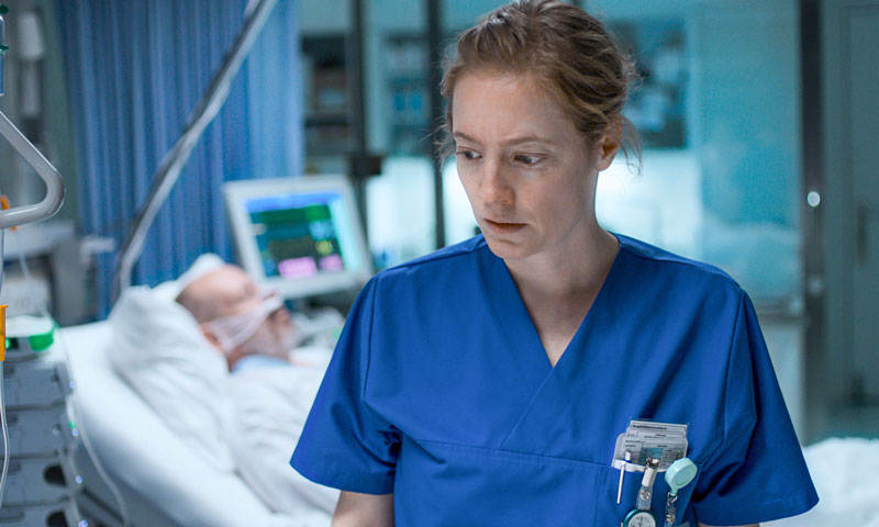 A nurse stands in front of the camera, with a patient in bed behind her, with a breathing aide and attached to a monitor 