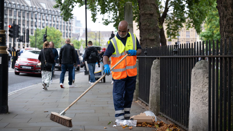 Abdul Jabbar with his broom, sweeping the street, with pedestrians walking in the background and a Parliament building in the distance 