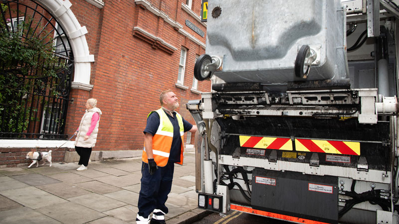 Ricky Rusby standing in the street next to a dustcart. A woman and her dog are in the background