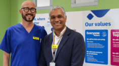 Baz Harding-Clark, dressed in hospital scrubs, and Alfredo Thompson in a suit, standing side by side and smiling. Behind them some information boards and the words 'our values'