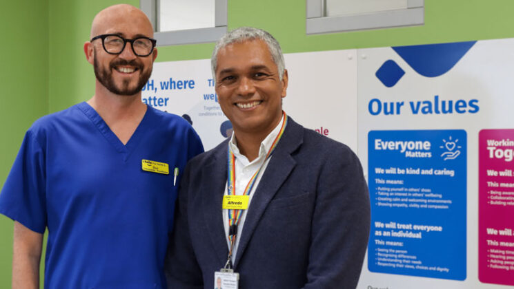 Baz Harding-Clark, dressed in hospital scrubs, and Alfredo Thompson in a suit, standing side by side and smiling. Behind them some information boards and the words 'our values'