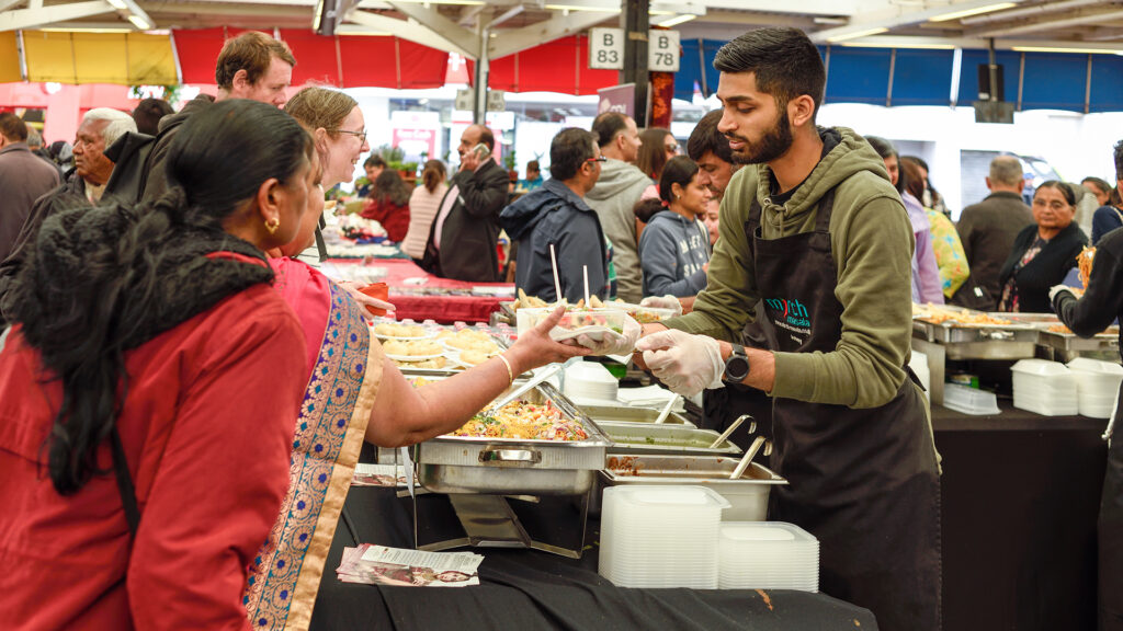 A BAME man serving two women some food in what looks like a busy market, with combination of white and BAME people in the background