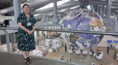 Tracey Wainwright standing on an upper level of what appears to be a conference centre, with people and stalls below, and a giant globe that takes up the whole height of the building