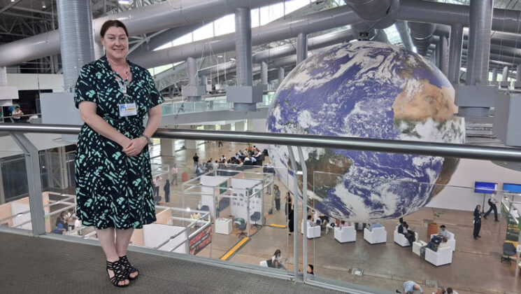 Tracey Wainwright standing on an upper level of what appears to be a conference centre, with people and stalls below, and a giant globe that takes up the whole height of the building