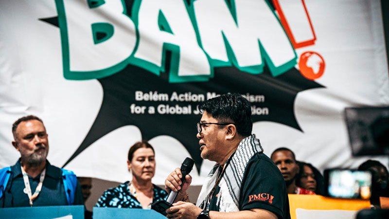 Ian Rivera of the Philippine Movement for Climate Justice, speaking with a microphone, as others listen. A BAM sign is in the background.