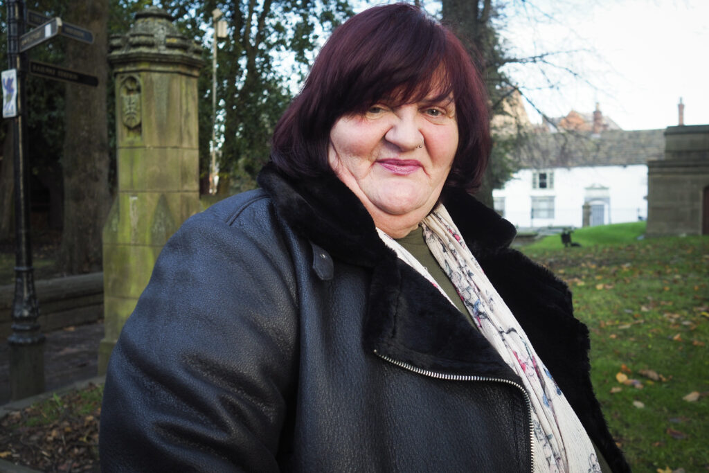Jeanette Lloyd is wearing a black jacket and looking into the camera. Behind her is a grassy area covered in autumn leaves