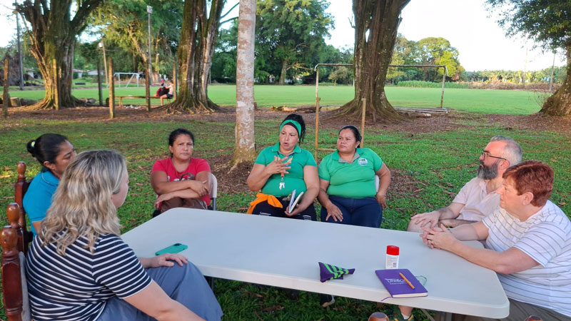 A group of workers and delegation members speaking around a table, in the open air. 