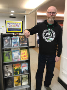 Alan Wylie, standing in a library alongside a feature stand containing books