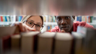 Photo from inside a book shelf, looking through the books at a man and a woman, who are looking in.
